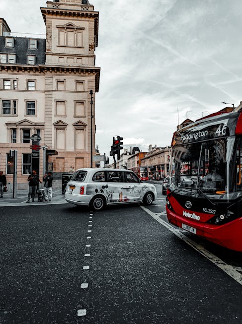 A street scene in Paddington showing a red and black double-decker bus with the route number 46, parked alongside a white taxi marked with advertising, both situated at a pedestrian crossing in front of an ornate beige building with multiple windows and decorative architectural details. The pavement is visible with pedestrians waiting to cross, some carrying bags, while the traffic light is red. The surrounding shops and buildings feature classic London architecture, and overcast skies cast diffuse light over the scene. This setting reflects typical urban logistics involved in house removals and packing and moving activities in Paddington, where vehicles like taxis and buses are parked near residential or commercial properties, and loading areas are often adjacent to busy streets.