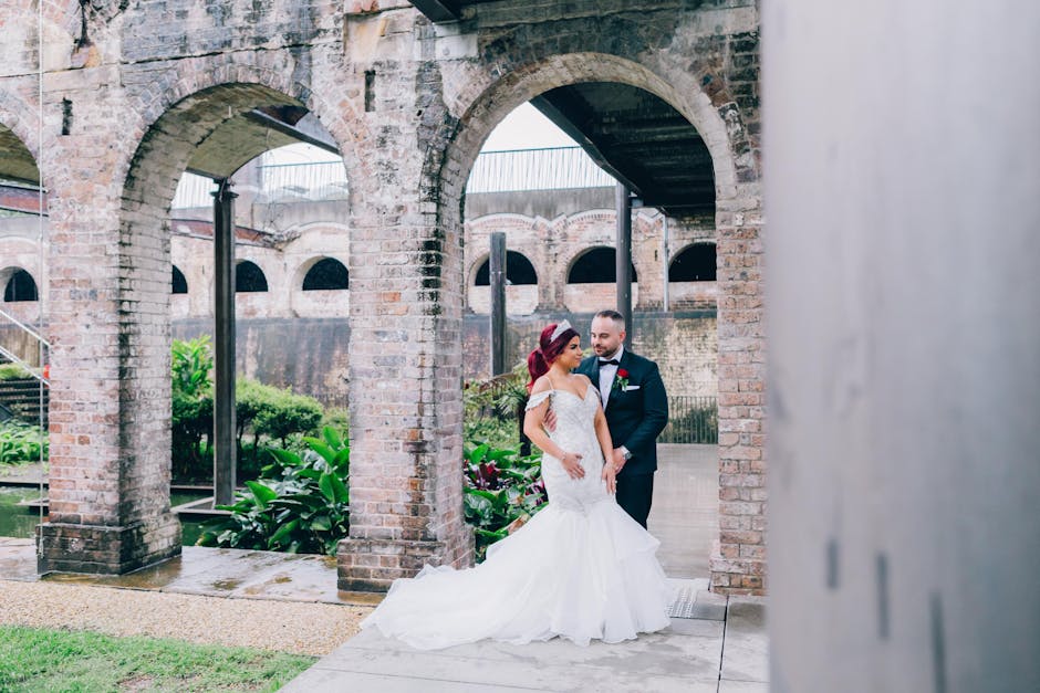 A bride in a white lace wedding gown and a groom in a dark suit with a white shirt and bow tie standing together under a brick archway outdoors. The bride has red hair styled in loose waves, and the groom has a beard and short hair. They are positioned on a concrete sidewalk, with lush green plants and flowers visible in the background. Behind them, there is a historic brick structure with multiple arches, some supporting a metal bridge or viaduct. The setting appears to be part of a historic site or garden with natural lighting illuminating the scene. This image captures a moment during a wedding photo shoot, with the couple standing close, engaged in conversation or looking at each other, amidst an environment featuring brickwork, greenery, and architectural details indicative of a heritage or outdoor venue.