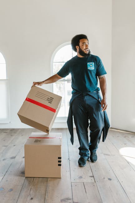 A professional mover with dark skin and an afro hairstyle is inside a bright, spacious room with white walls and a large arched window allowing natural daylight. He is dressed in a navy blue uniform with a company logo on the chest and has a black jacket tied around his waist. The mover is holding a cardboard box wrapped with red tape in his right hand, preparing to place it onto a second cardboard box positioned on the wooden floor. The boxes are standard packing containers used in home relocation, with the floor featuring light-colored wooden planks. The movement appears to be part of a packing and loading process typical of house removals, with no furniture or other items visible, focusing solely on the packaging materials and the interior setting. The scene subtly reflects the logistics involved in furniture transport and home relocation services provided by Removal Companies Paddington.