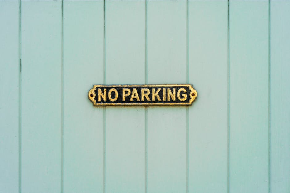 A rectangular sign with a black background and raised gold lettering displaying 'NO PARKING' is mounted on a light green wooden wall comprised of vertical panels. The sign appears to be made of metal, with visible mounting brackets on each side. The wall may be part of a residential or commercial building in a parking-restricted area. The image is taken during daylight, with even lighting highlighting the texture of the wooden panels and the sign's details. This visual context supports the understanding of parking restrictions, which can be relevant for house removals and furniture transport when navigating tight or restricted access points in W2, Paddington. Removal Companies Paddington may encounter such signage during the home relocation process, especially when coordinating parking or delivery access for moving services.