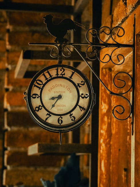 A vintage-style round clock with a white face and black numerals is mounted on an ornate black metal bracket attached to a brick wall. The clock displays the time as approximately 8:11 and bears the inscription 'Paddington Station, London, 1854' on its face. Above the clock, a decorative black metal silhouette of a dog sits atop the bracket. In the background, a wooden staircase with visible steps and a handrail can be seen, illuminated by warm lighting, suggesting an indoor setting. The scene captures a nostalgic, traditional interior atmosphere, relevant to home relocations and moving services provided by Removal Companies Paddington, emphasizing the importance of timing and access during house removals near Paddington Station.