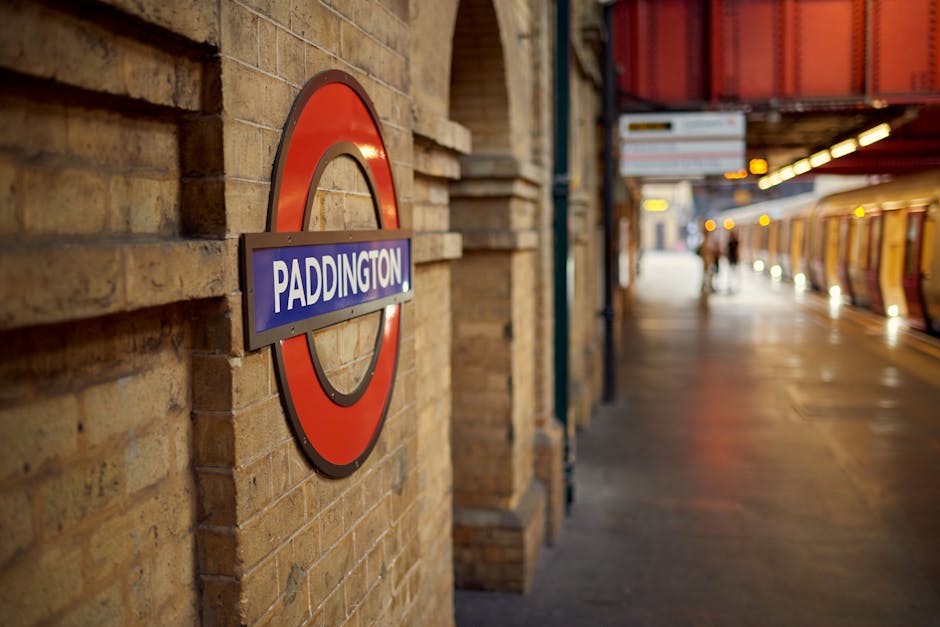 Close-up view of the Paddington underground station sign mounted on a light brown brick wall near the platform, with the station's distinctive red, blue, and black circular design and white text reading 'Paddington'. In the background, the platform extends with a row of benches and commuters waiting, illuminated by station lighting. To the right, a partially visible orange and cream train is stationary, with closed doors and a few passengers walking along the platform. The scene captures the typical busy atmosphere of London transport hubs, relevant to the context of home relocation and furniture transport services offered by Removal Companies Paddington, who handle loading and unloading processes at such stations during moves.