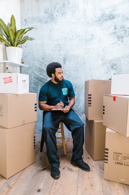 A man with dark skin, curly black hair, and a beard, dressed in a teal T-shirt and dark pants, is seated on a wooden stool inside a room with a textured light blue wall. He is holding a clipboard and pen, appearing to be taking notes or conducting an inventory during a house or office relocation. Surrounding him are several cardboard moving boxes of various sizes, some sealed with packing tape and others open, stacked on the wooden floor. To his left, there is a white shelving unit with a large potted plant with green, spiky leaves and a white box labeled 'GLASS' on the top shelf. The scene suggests the process of packing or managing items for a removal service, with natural lighting illuminating the space, reflecting typical packaging and organization activities associated with professional removals that [COMPANY_NAME] might provide as part of their moving and furniture transport services. The setting emphasizes careful handling and preparation for a home or office move, consistent with the context of house removals and moving logistics.