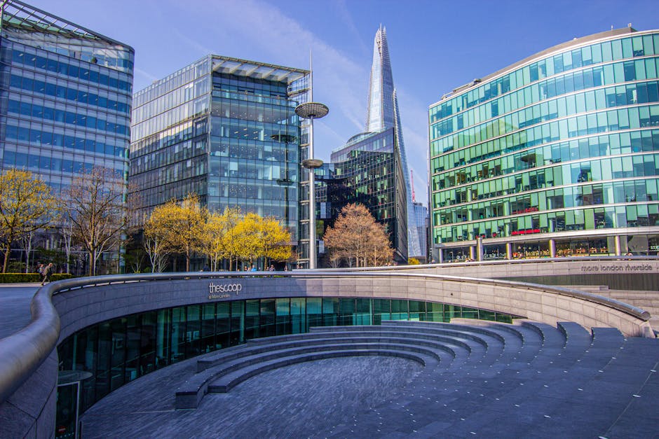 A man with dark skin, curly black hair, and a beard, dressed in a teal T-shirt and dark pants, is seated on a wooden stool inside a room with a textured light blue wall. He is holding a clipboard and pen, appearing to be taking notes or conducting an inventory during a house or office relocation. Surrounding him are several cardboard moving boxes of various sizes, some sealed with packing tape and others open, stacked on the wooden floor. To his left, there is a white shelving unit with a large potted plant with green, spiky leaves and a white box labeled 'GLASS' on the top shelf. The scene suggests the process of packing or managing items for a removal service, with natural lighting illuminating the space, reflecting typical packaging and organization activities associated with professional removals that [COMPANY_NAME] might provide as part of their moving and furniture transport services. The setting emphasizes careful handling and preparation for a home or office move, consistent with the context of house removals and moving logistics.