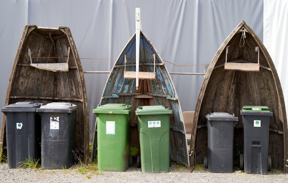Four small wooden boats, turned upside down, are positioned side by side outdoors, with the open interiors facing upward. Behind each boat, there are vertical lines of orange string or rope, possibly used for securing or drying. In front of the boats, there are five wheeled rubbish bins—two black, two green, and one dark grey—with closed lids, placed on a gravel surface. Some of the bins have labels or stickers. The scene appears to be in a sheltered area with a light-colored fabric or plastic sheet in the background, suggesting a temporary setup. The context relates to house removals and packing, where boats may be stored or transported as part of furniture or equipment during a home relocation, with general waste and recycling bins ready for disposal or collection managed by Removal Companies Paddington, who specialise in removals and moving logistics.