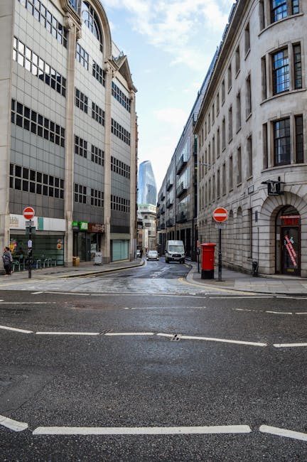 A city street scene showing a narrow road with asphalt pavement and white road markings, flanked by tall, multi-story office buildings made of concrete and glass on both sides. The buildings have numerous windows, some with reflective glass. On the left side, there is a small green storefront and a pedestrian walking nearby, while on the right side, a red telephone booth is situated against the building wall. The street features a few parked vehicles, including a white van, and a red mailbox is positioned near the telephone booth. The sky above is partly cloudy, and the scene suggests an urban environment suitable for house removals and furniture transportation, with space for loading and unloading activities. The image supports understanding of logistical considerations involved in home relocation within city streets, highlighting typical urban street closures, loading bays, and parking regulations managed by Removal Companies Paddington.