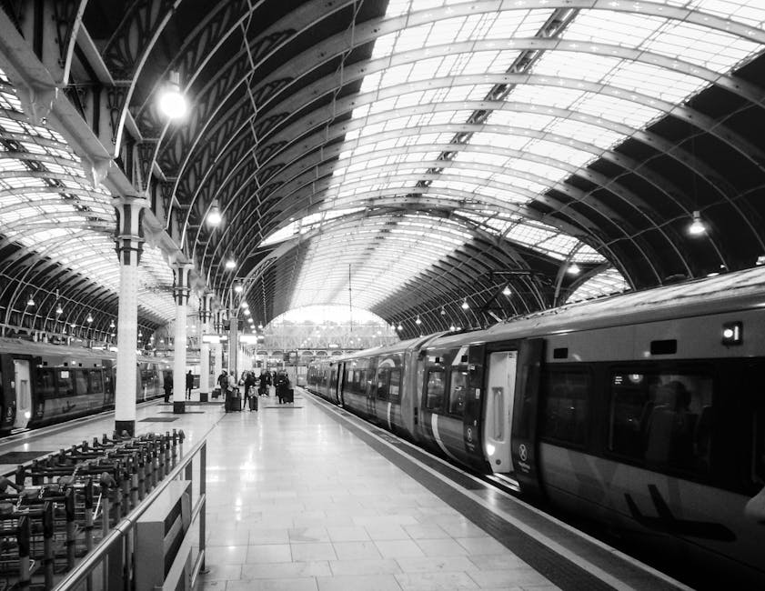 Inside a spacious railway station with a high, arched glass and metal ceiling illuminated by bright lights. In the foreground, a modern yellow and black train is positioned on the tracks, with red loa