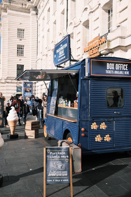 A blue mobile food cart is situated on a busy city street with a large group of people gathered around. The cart has a counter for serving and is decorated with images of popcorn, indicating it offers snacks or treats. An A-frame sign in front of the cart displays information about ticket purchases, suggesting it is part of a public event or entertainment setup. Behind the cart, there are tall, white stone buildings with multiple windows, typical of urban architecture. To the left, there is a large ice cream sculpture and a stack of wooden crates, potentially used for storage or delivery purposes. The scene appears well-lit with natural daylight, and the environment includes pedestrians walking and standing, some engaged in conversation or waiting. This setting relates to house removal services, as such setups are common at moving or event locations, where removals companies like Removal Companies Paddington may provide logistical support for relocations or related services.