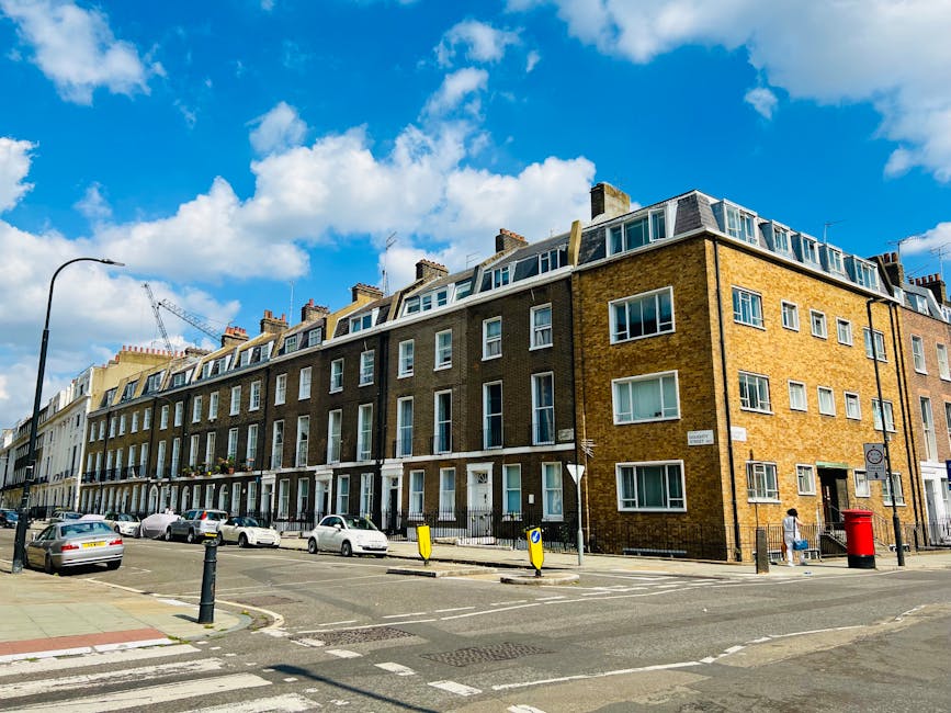 A wide view of a multi-storey residential building on a city street under a blue sky with scattered white clouds. The building has a brown brick exterior with large, white-framed windows and dormer windows on the top floor, indicating attic conversions. The street in front features parked cars, a pedestrian walking along the sidewalk, and various street furniture including a black lamppost, a yellow and black bollard, and a red post box. Near the building's entrance, there are visible moving boxes and furniture covered with protective blankets, suggesting a home relocation. In the foreground, a loading area with damaged pavement is present, and a crane is partially visible in the background, indicating ongoing construction or renovation work. The scene reflects an urban environment suited for professional removals and house moving services, as offered by Removal Companies Paddington.