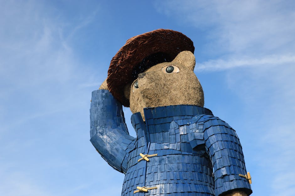 A large outdoor sculpture of a human figure with a textured, stone-like face and a large brown hairstyle, wearing a blue garment made of small, overlapping rectangular panels. The figure is positioned against a clear blue sky, with its left hand raised, touching the head. The sculpture appears to be part of a public art installation. This image is relevant to house removals and packing services provided by Removal Companies Paddington, as it demonstrates the importance of careful handling and transport of large, fragile objects during home relocation and furniture transport processes.