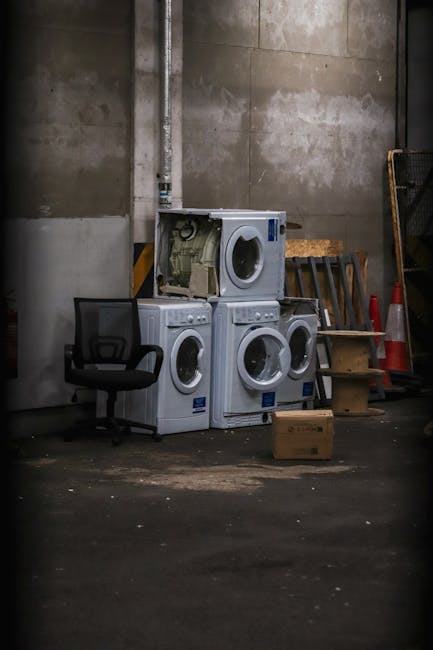 A set of four front-loading washing machines and dryers stacked in a storage area with exposed concrete walls and industrial piping. The appliances are white, with one dryer door open revealing internal components. To the left, there is a black office chair without armrests placed next to the appliances. Behind the appliances, a wooden chair with a slatted back and seat is visible, alongside a small wooden platform or step. On the right, orange and white traffic cones are arranged against the wall. The floor is dark and appears to be concrete, with some dust or debris. The scene suggests a space used for storing appliances and equipment awaiting disposal, recycling, or preparation for a home relocation or packing and moving service, typical in the context of managing bulky waste after a Paddington move, as handled by Removal Companies Paddington.