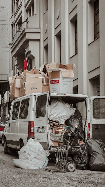 A white removal van parked on a city street next to a multi-storey building with classic architectural features. The van’s rear doors are open, revealing large black plastic trash bags, flattened cardboard boxes, and other packaging materials stuffed inside. On top of the van, numerous cardboard boxes of various sizes, some partially crushed, are stacked, secured with packing tape and plastic wrap. A small hand truck is positioned at the back of the van, loaded with more black bags and flattened boxes, indicating a packing and loading process typical of home relocation efforts. The scene suggests the disposal or temporary storage of bulky waste, possibly as part of packing down items during a removal service carried out by companies like Removal Companies Paddington. The environment features a neutral tone with muted lighting, emphasizing the practical aspect of managing waste after a move.